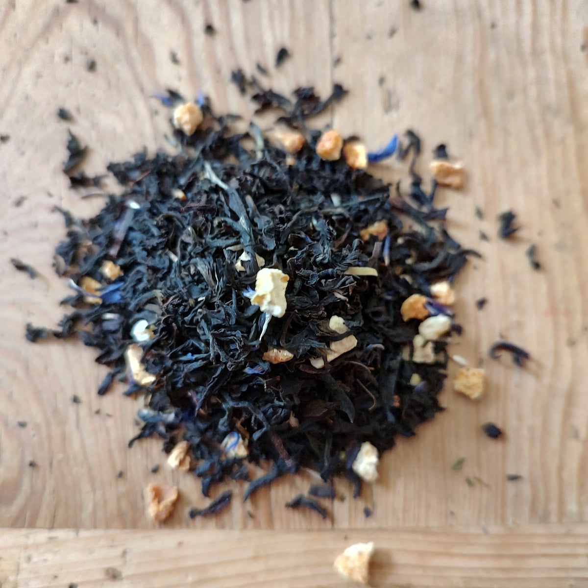 A small pile of loose-leaf tea on a light wood surface, featuring dark black tea leaves with small beige dried fruit pieces and a few blue dried flower accents.