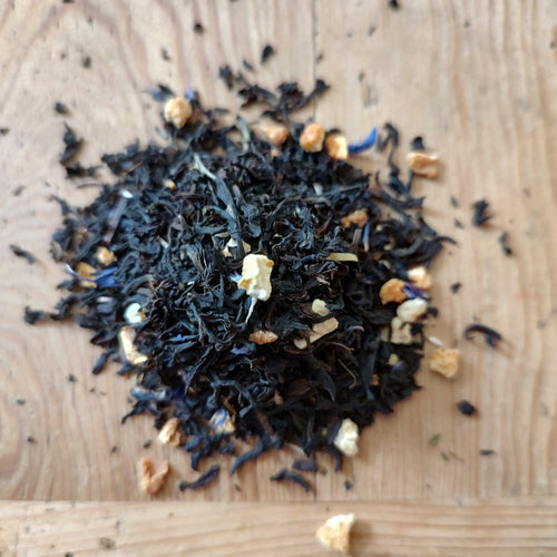 A small pile of loose-leaf tea on a light wood surface, featuring dark black tea leaves with small beige dried fruit pieces and a few blue dried flower accents.