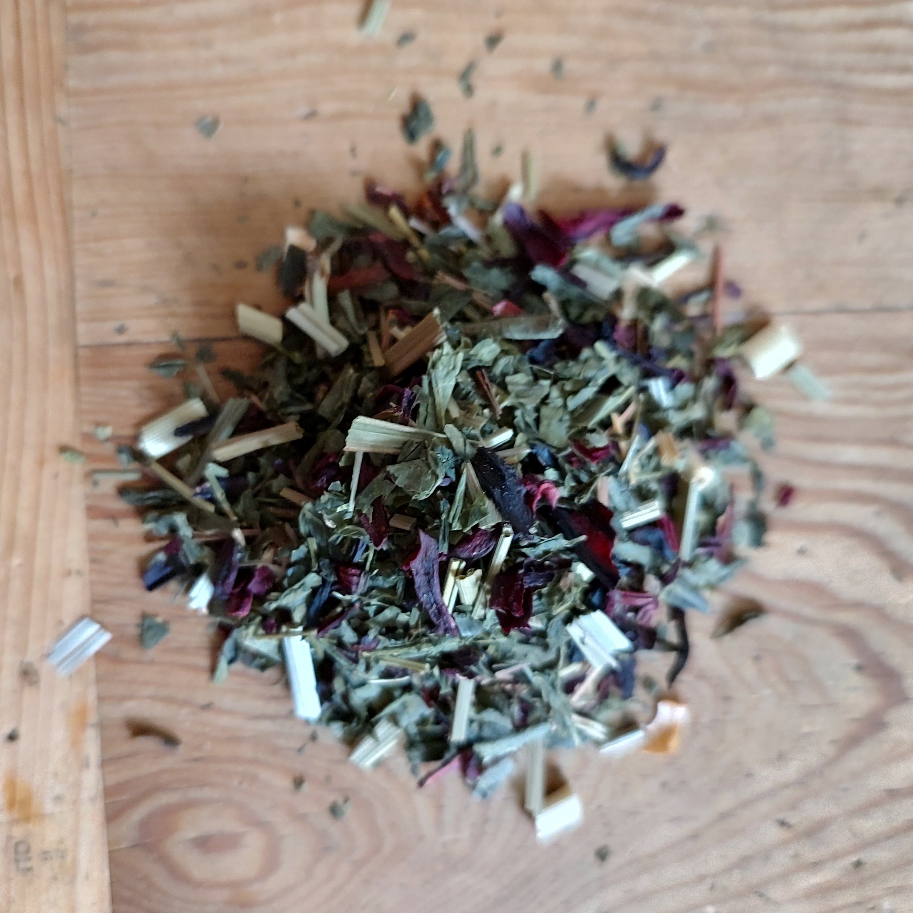 A small pile of loose-leaf herbal tea blend containing dried green, brown, and purple botanicals, scattered on a light-colored wooden surface.