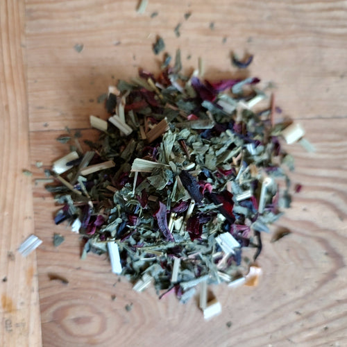 A small pile of loose-leaf herbal tea blend containing dried green, brown, and purple botanicals, scattered on a light-colored wooden surface.