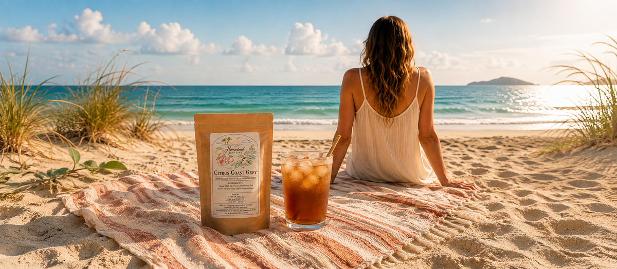 A woman sits on a striped beach towel on the sand facing the ocean, with a stand-up pouch and an iced coffee drink in front of her. The scene is set on a sandy beach with dune grass and a blue sky.
