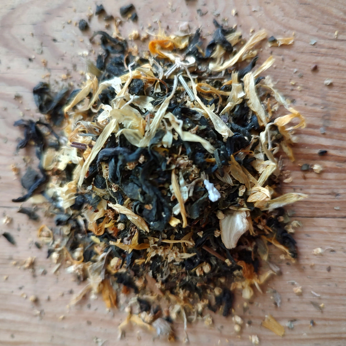 A small pile of loose-leaf herbal tea or spice blend containing dark dried leaves, light tan pieces, and small white bits, placed on a wooden surface.