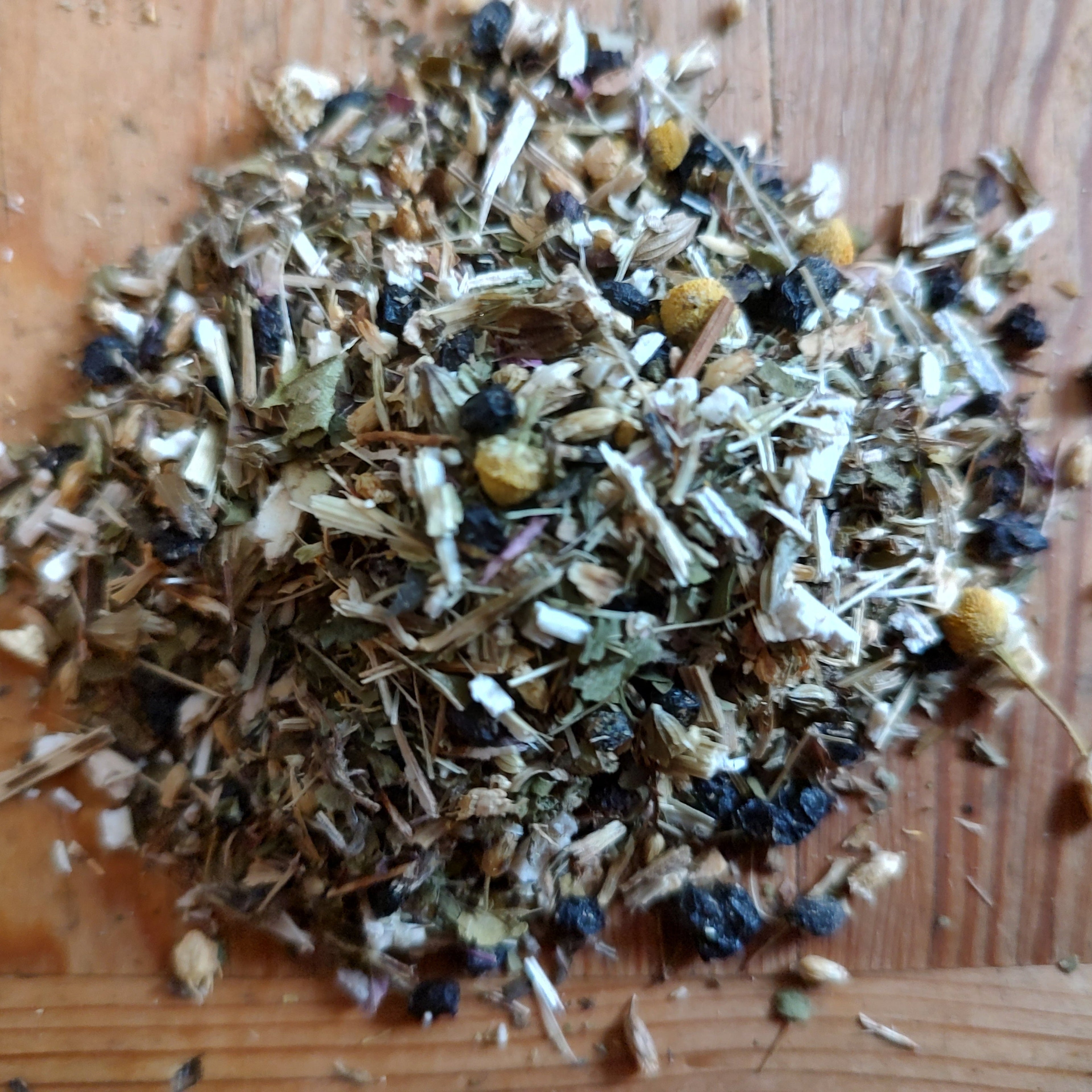 A pile of loose-leaf herbal tea or spice blend containing dried flowers, leaves, seeds, and bark pieces, elderberries, displayed on a wooden surface.