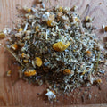 A pile of loose-leaf herbal tea or tisane made from dried chamomile flowers and green leaves, resting on a wooden surface.