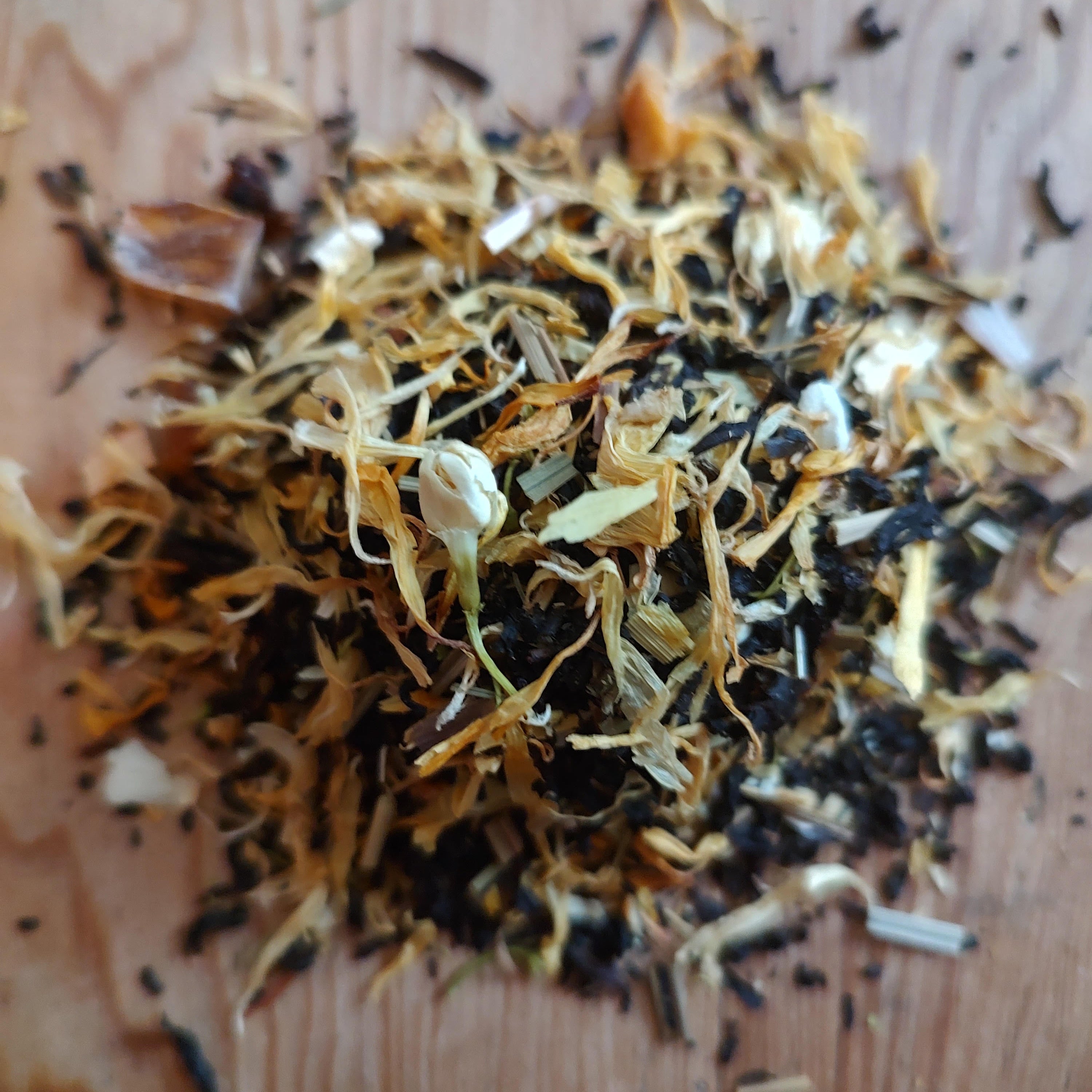A pile of loose-leaf herbal tea or spice blend containing dark brown leaves, golden-brown dried flowers, and small white blossoms, displayed on a light-colored wooden surface.