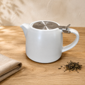 A white ceramic teapot with a stainless steel infuser on the lid, set on a light wooden table next to a folded beige cloth and loose tea leaves.