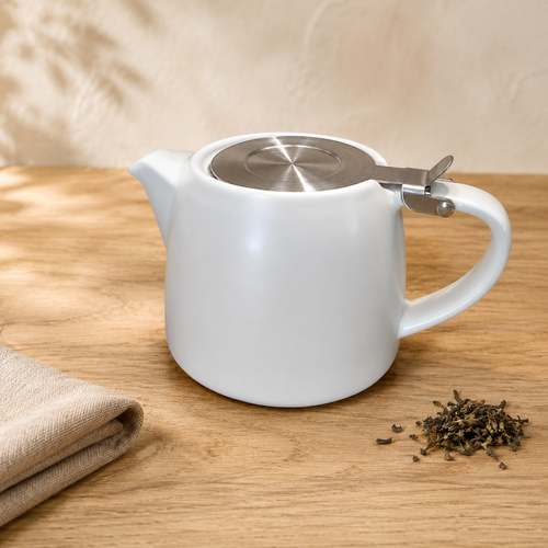 A white ceramic teapot with a stainless steel infuser on the lid, set on a light wooden table next to a folded beige cloth and loose tea leaves.